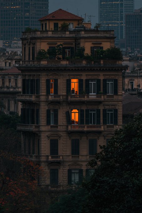 A moody twilight photograph of a classic multi-story European building, with warm light glowing from two windows where silhouetted figures are visible against a backdrop of a dark city skyline.