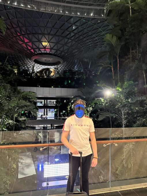 A person wearing a white 'RICHMOND' t-shirt and a glowing LED mask pulling a suitcase inside a large indoor venue with tropical greenery and a distinctive domed glass ceiling, resembling Jewel Changi Airport.