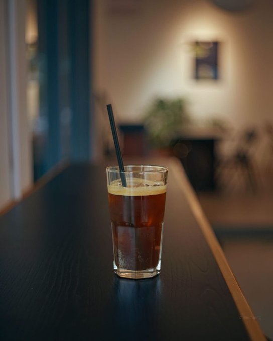A tall glass of iced coffee with a black straw sits on a dark wooden counter, with a softly blurred background showing a plant and framed artwork.
