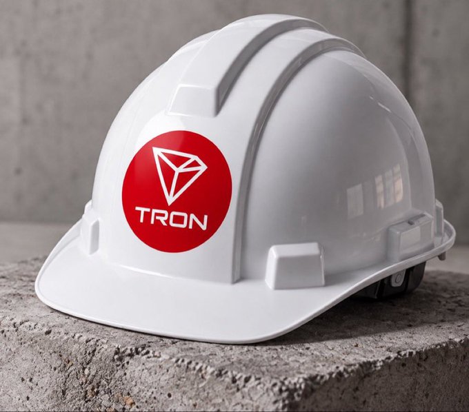 A white hard hat sitting on a concrete ledge, featuring a red circular logo with a diamond/geometric symbol and the word 'TRON'.