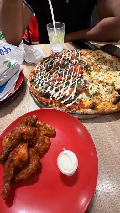 A table set with a large pizza, a red plate of chicken wings with dipping sauce, and a glass with a straw.