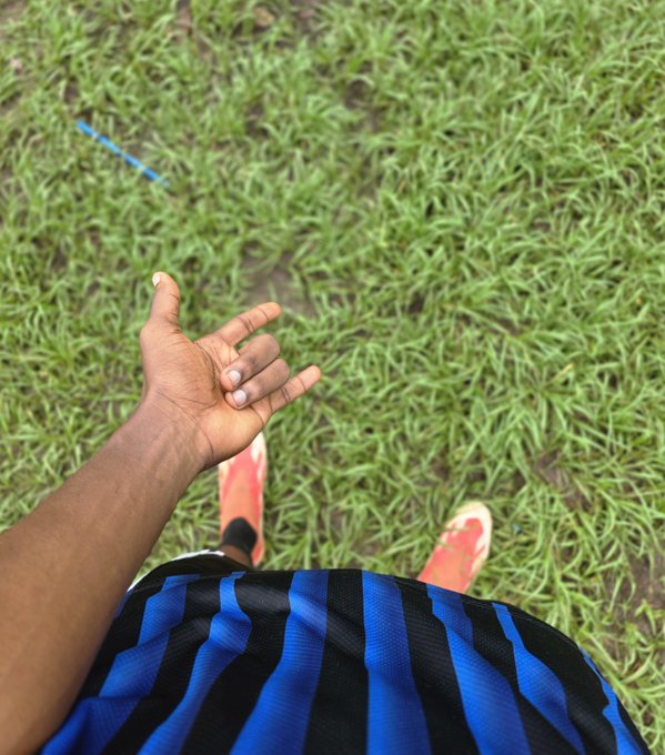 A first-person perspective looking down at a person's hand extended outward, wearing a blue and black striped shirt and red shoes, standing on a patch of green grass.
