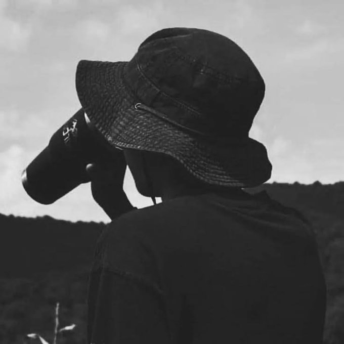A black and white photo of a person viewed from behind, wearing a bucket hat and holding a camera up to their face.