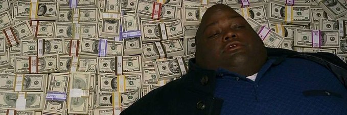 A man in a blue shirt lies amid a massive pile of bundled U.S. dollar bills.