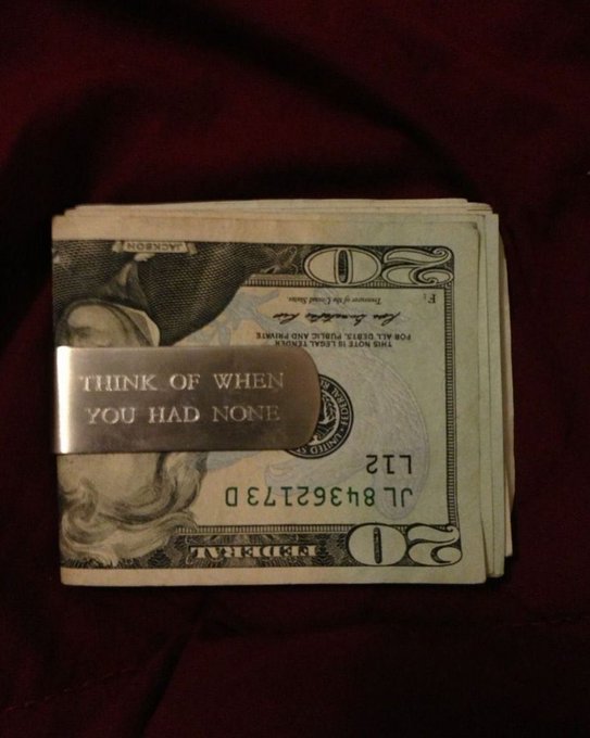 A stack of U.S. twenty-dollar bills held together by a metal money clip engraved with 'THINK OF WHEN YOU HAD NONE', resting on dark red fabric.
