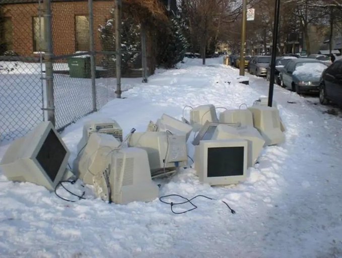 A pile of discarded CRT monitors sits abandoned on a snow-covered sidewalk next to a chain-link fence.