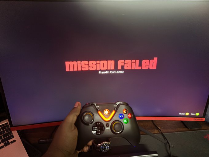 A hand holds an Xbox-style controller in front of a TV displaying "Mission Failed" in red text from the game Saints Row, while a laptop and watch sit on a desk nearby. The "Mission Failed" screen humorously mirrors the poster's feeling of failing at life—from being unemployed and drained by crypto ($POLY) investments to lacking a girlfriend and a PS5 for GTA VI.