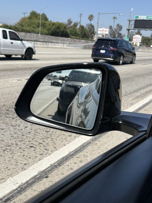 A view from inside a car showing the side mirror reflecting the dark vehicle's body, with traffic and a highway overpass visible on the road ahead.