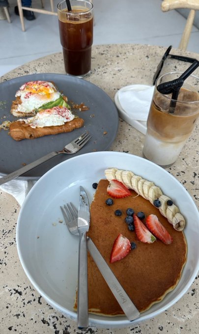 A cafe table with a large pancake topped with strawberries, banana, and blueberries on a white plate, accompanied by an iced latte and a savory dish with avocado and eggs.