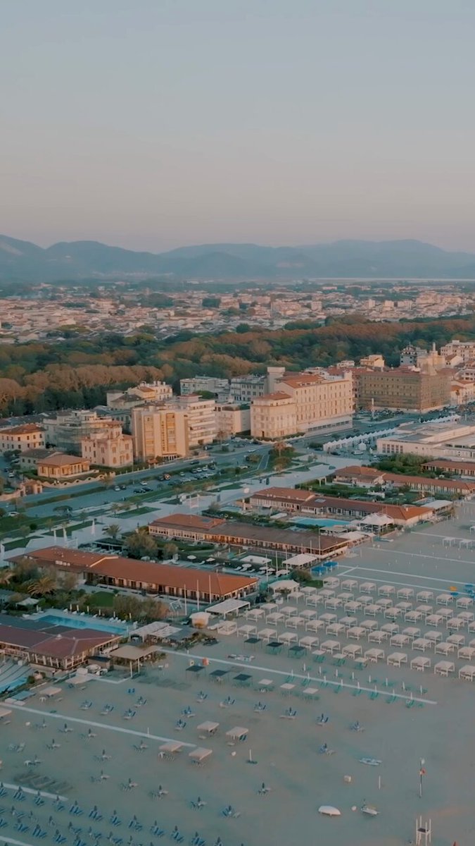 The image features an aerial view of a city with a beachfront area below. The cityscape includes buildings, roads, and a sandy beach. There are numerous umbrellas scattered throughout the scene, indicating that this location is likely a popular spot for relaxation or leisure activities. The presence of Bitcoin in the image suggests that the city may be associated with cryptocurrency or digital currency markets.