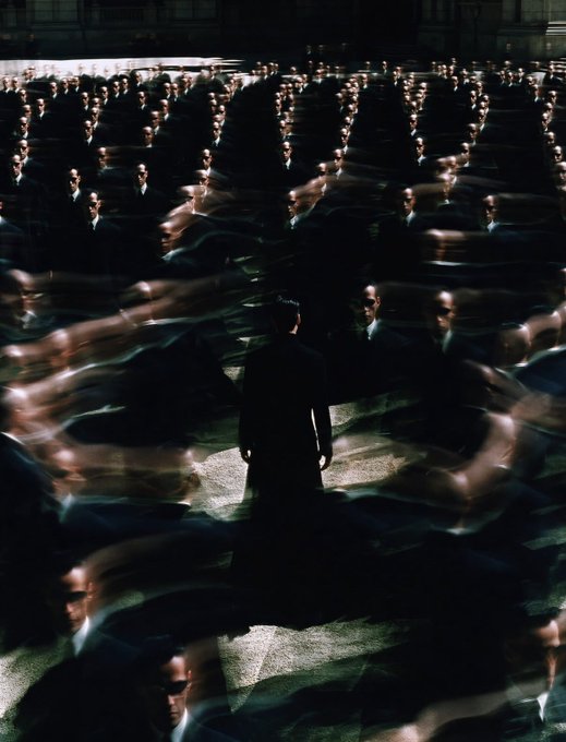 A dark cinematic image showing a solitary figure in a suit surrounded by rows of identical men in suits, resembling the Agent Smith scene from The Matrix.