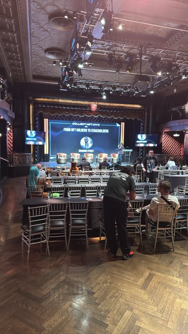 A photograph taken from the back of a banquet hall set up for a poker tournament, with a large screen displaying 'COLLATERAL NFT 10/10 FROM NFT FAILURES TO FRAUDULATORS' and several players seated at round tables.