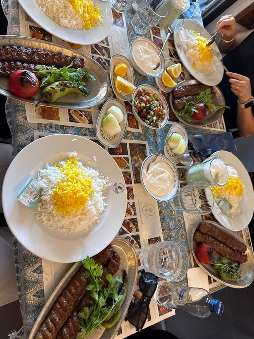 A top-down view of a dining table covered with plates of white and yellow rice, kebabs, grilled vegetables, and various Middle Eastern side dishes like salad and yogurt.