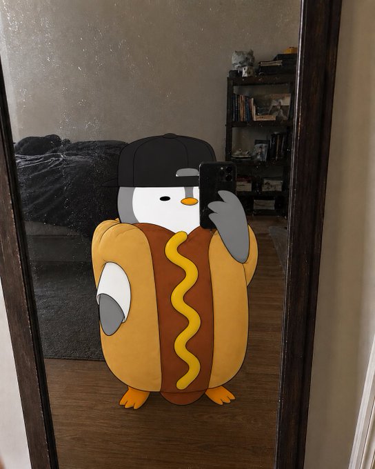 A mirror selfie showing a person wearing a digitally-added penguin costume dressed as a hot dog, complete with a black baseball cap, standing in a bedroom.