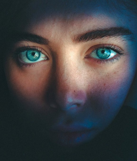 A close-up portrait of a person's face, focusing on striking blue-green eyes illuminated against a dark, moody background with dramatic blue lighting.