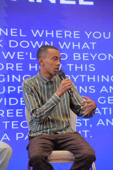 A man wearing a green and orange striped shirt sits on a chair holding a microphone, speaking in front of a bright blue backdrop with partial white text visible.