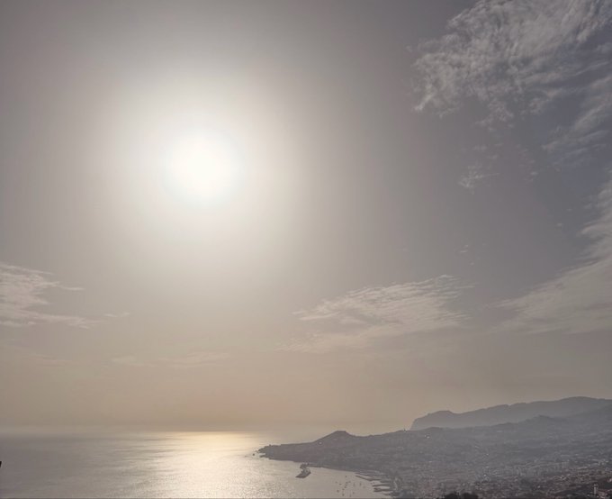 A scenic view of a bright sun shining over a calm sea with a coastal city and mountains visible on the horizon.
