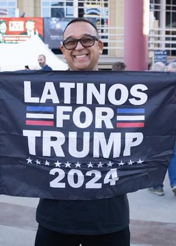 A man smiles while holding a black "LATINOS FOR TRUMP 2024" banner with red and blue accents and stars, directly visualizing the demographic shift referenced in the post about Latino men swaying between Republican and Democratic support.