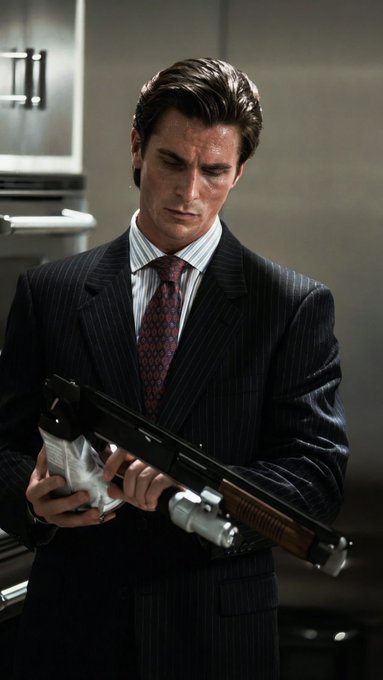 A man in a dark pinstripe suit and patterned tie holds a shotgun and a wrapped package, looking downward in what appears to be a kitchen setting.