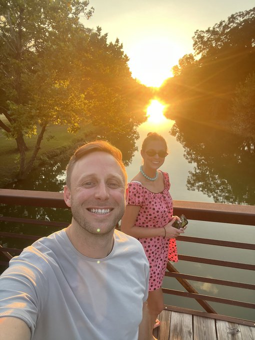 A smiling couple takes a sunset selfie on a wooden deck overlooking a calm river, with the man holding the camera and the woman in a pink polka-dot dress behind him. The warm, glowing sunset and relaxed setting perfectly capture the laid-back vibe of a "Solid Saturday."