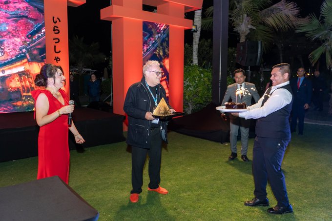 An evening outdoor event with Japanese signage ('いらっしゃ') where a woman in a red dress holds a microphone, while a man in glasses receives a golden tray from a server.