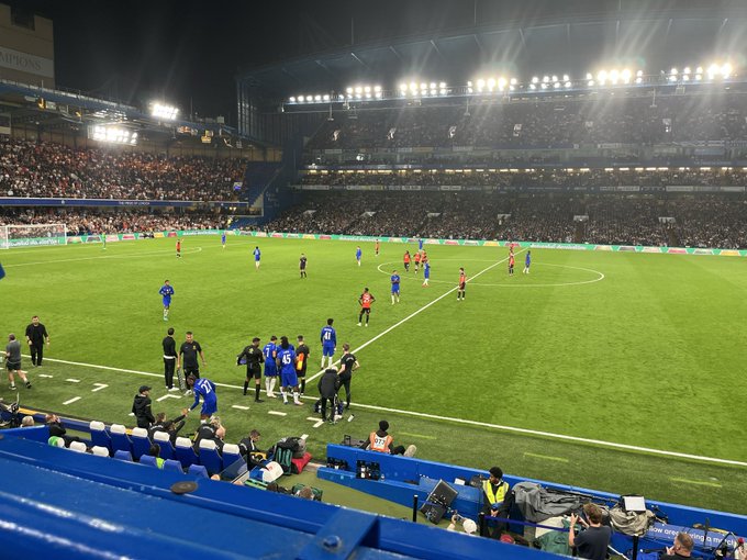 A nighttime view of a soccer match in a large stadium with bright floodlights, showing players in blue and red kits on the field, with a crowded spectator stand and media personnel in the foreground.