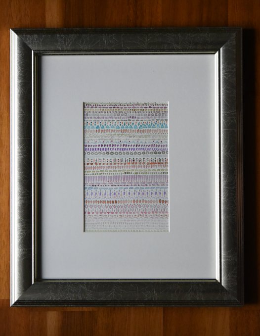 A framed piece of textile art displayed on a wooden surface, featuring rows of colorful hand-stitched patterns in pastel tones within a distressed silver frame.