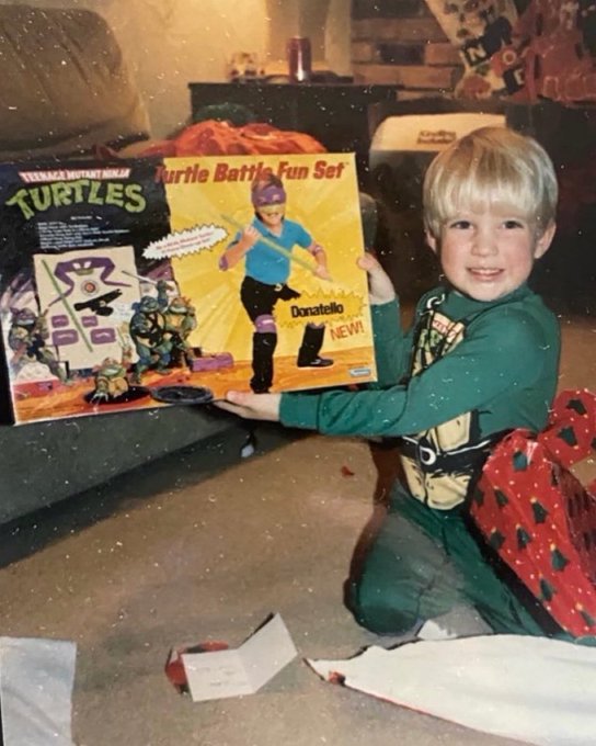 A young blonde child kneels on the floor smiling while proudly holding up a vintage "Teenage Mutant Ninja Turtles Turtle Battle Fun Set" featuring Donatello, with a heartfelt caption reflecting on their lifelong love for the TMNT character and excitement for an upcoming VeVe digital collectible drop on July 4th.