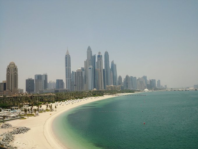 A daytime view of a coastal city skyline with numerous skyscrapers rising behind a sandy beach and turquoise water.
