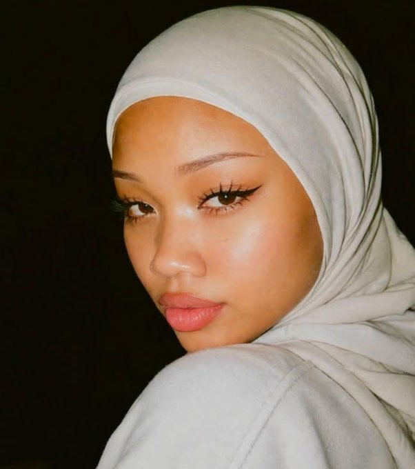 A close-up portrait of a woman wearing a light gray hijab, featuring defined eyebrows, winged eyeliner, and glossy pink lips against a dark background.