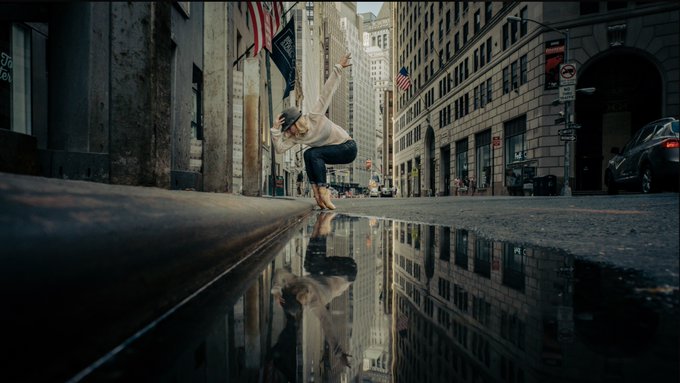A woman in a hat and white top balances playfully on the edge of a puddle on a city street, her reflection mirrored in the water below as tall buildings and an American flag rise in the background.