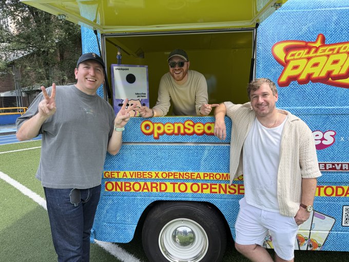 Three cheerful men pose by a bright blue "Collector Park" van featuring OpenSea branding and text promoting "Get a Vibes x OpenSea Promo Onboard to OpenSea," with one man leaning out of the service window holding a Mark Rober product, capturing the lively, promotional spirit of the event.