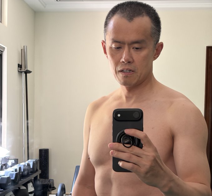 A shirtless man with short hair takes a mirror selfie in a gym using a smartphone with a grip attachment, showing gym equipment in the background.