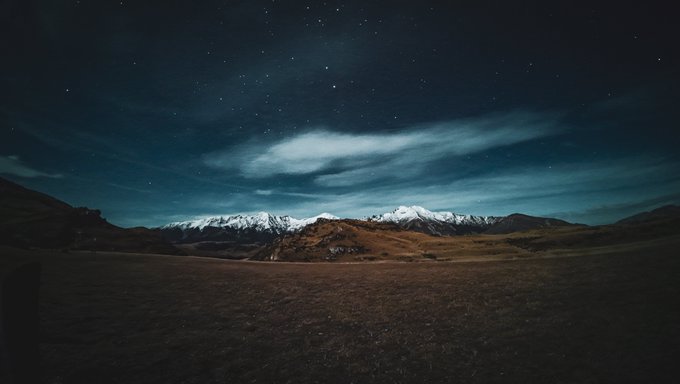 A nighttime landscape shows snow-capped mountains illuminated under a dark sky filled with stars and wispy clouds, with brown grassy hills in the foreground.