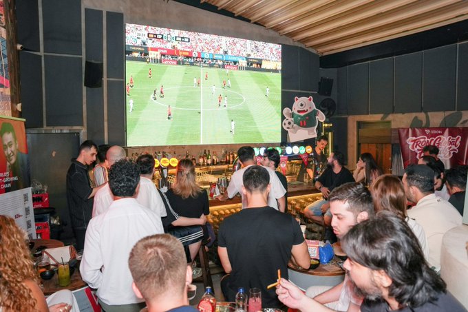 A photograph of a crowded bar where people are gathered to watch a soccer match on a large projection screen, with a Budweiser banner visible on the right.