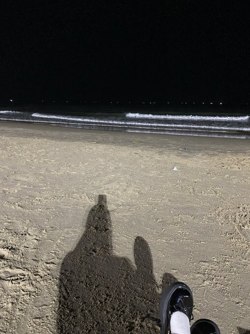 A nighttime beach scene showing the shadow of a person sitting in the sand, with a sneaker and white sock visible in the foreground, and the ocean in the background.