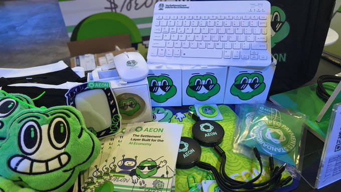 A promotional display featuring AEON-branded merchandise, including a green plush mascot, white wireless keyboard and mouse, lanyards, stickers, and a flyer reading "The Settlement Layer Built for the AI Economy," all arranged at the AEON booth PB1-13 at TOKEN2049 Singapore.