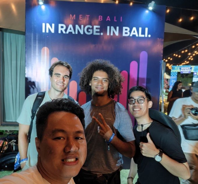Group of friends posing for a selfie in front of a banner reading 'MET BALI IN RANGE. IN BALI.'