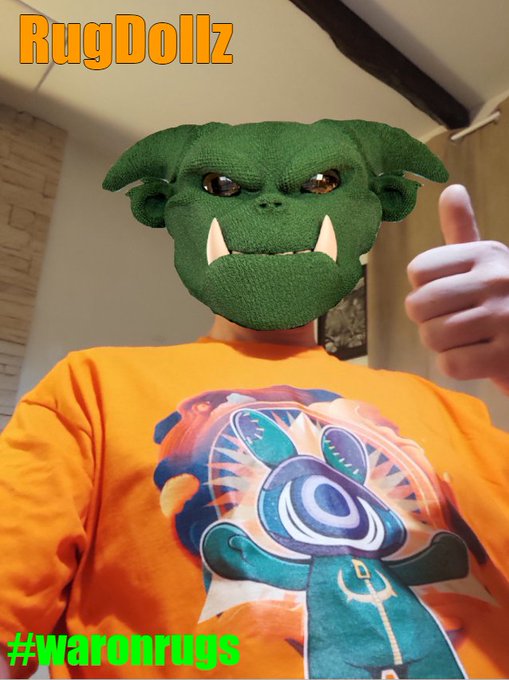 A person wearing a green troll/orc mask with tusks and an orange t-shirt featuring a cartoon character gives a thumbs up, with text reading 'RugDollz' and '#waronrugs'.