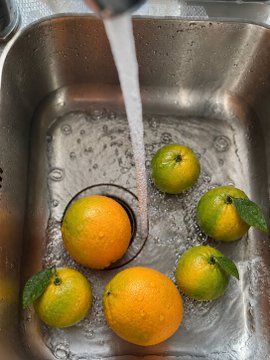 Several oranges and green citrus fruits being washed under running water in a stainless steel sink.