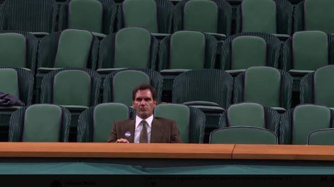 A scene of a man in a brown suit and tie sitting alone in empty green stadium seats at Wimbledon, with a nameplate reading 'The Hon. Petchey' partially visible at the bottom.