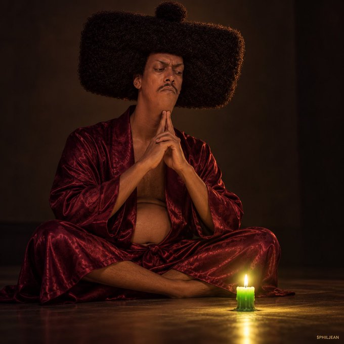 A man with a large afro wearing a red velvet robe sits cross-legged in meditation beside a lit candle, with a watermark reading '$PHILJEAN'.