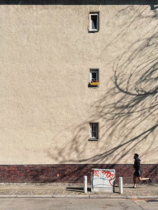The image shows an urban street scene with a tall building, a person running down the sidewalk, and a sign that reads "GM (not a real gm until Iran is free) & happy almost Friday!!! Thrilled to see that @SofieUgly outbid @328_lad on @55 XTZ now and you can place a bid for only 1 XTZ (30 cents) more". The person running down the sidewalk appears to be in motion, possibly enjoying their run or rushing to reach an important destination.