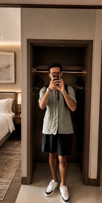 A man wearing a light green button-up shirt, black shorts, and white sneakers takes a mirror selfie with his phone while standing in a hotel room closet, with a bed visible in the background.