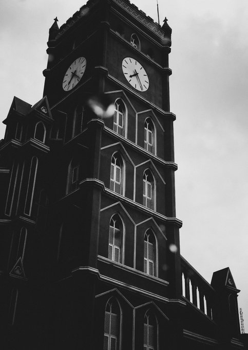 A black-and-white photograph looking up at a tall, gothic-style clock tower with two visible clock faces, featuring arched windows and dark architectural details against a cloudy sky.