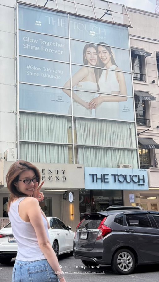 A woman in a white tank top and glasses taking a selfie with a peace sign in front of a large building advertisement for 'THE TOUCH CLINIC' featuring two models and the text 'Glow Together Shine Forever'.