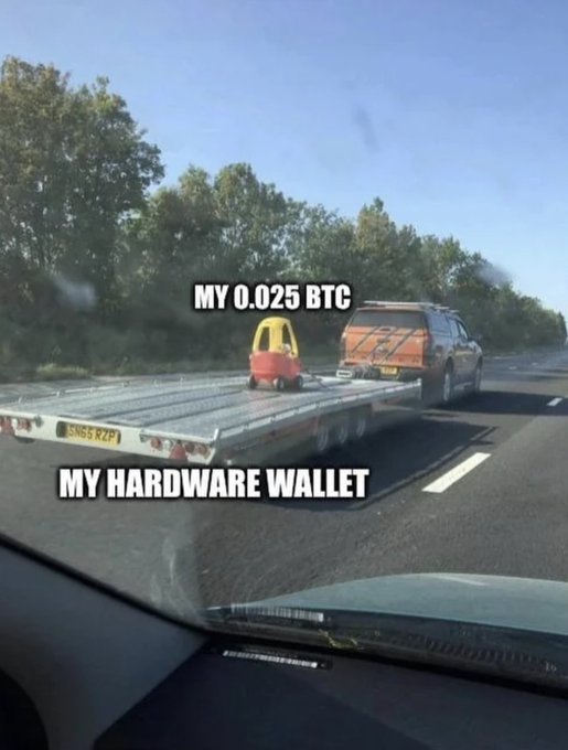 A view from inside a car shows a flatbed tow truck on the highway carrying a small Little Tikes toy car on its back. Text overlays label the toy car as "MY 0.025 BTC" and the flatbed tow truck as "MY HARDWARE WALLET," humorously comparing the tiny toy vehicle to a valuable crypto asset.