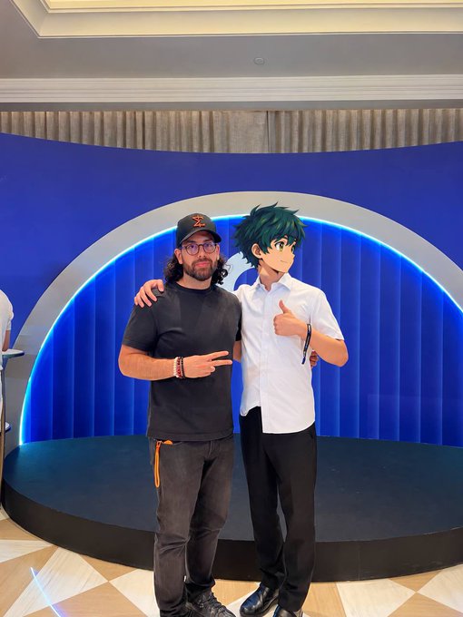A bearded man in a black cap and t-shirt poses alongside a person dressed as the anime character Izuku Midoriya from *My Hero Academia*, both flashing peace and thumbs-up signs at what appears to be the Gate Gala event. The setting, a sleek blue-lit stage with a circular arch backdrop, aligns with the post's mention of catching up with old friends at the @Gate Gala while discussing @0xMiden Mainnet developments.