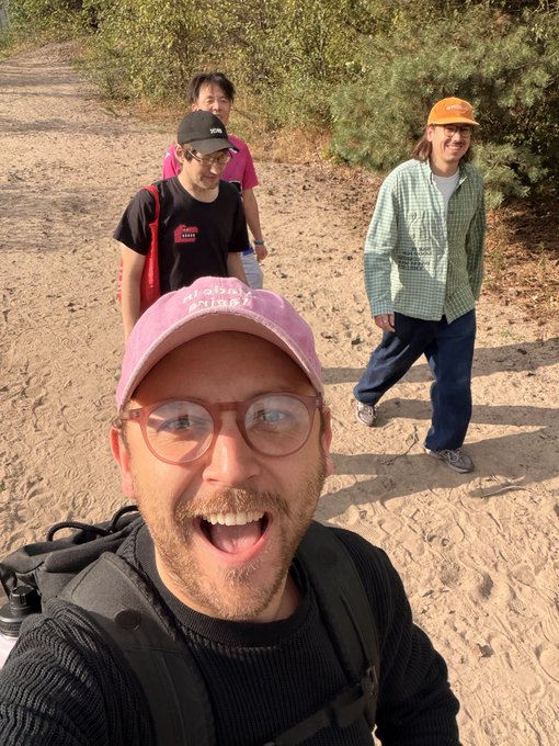 A selfie of a man wearing a pink 'Anatomy of a Murder' cap and glasses, grinning widely, with three other people walking behind him on a sandy path surrounded by greenery.