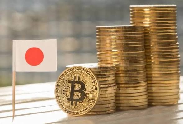 A small Japanese flag stands next to a Bitcoin coin and stacks of gold coins, suggesting a connection between Japan and cryptocurrency investment.